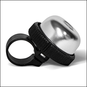 A imagem mostra uma campainha de bicicleta redonda na cor prata com um apoio preto em sua base. O fundo da imagem é branco.