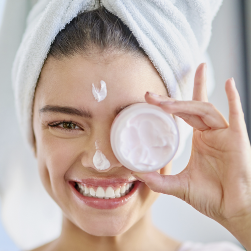 Imagem de uma mulher caucasiana com traços orientais sorrindo, com uma toalha na cabeça e segurando um creme facial na mão esquerda.