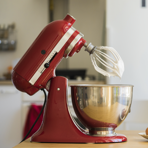 Imagem de uma batedeira vermelha, modelo Artisan da Kitchenaid, com sua cabeça levantada mostrando os batedores sujos de clara em neve.