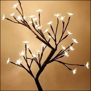 A imagem mostra uma árvore de cerejeira com os galhos pretos e as flores brancas com pequenas luzes de led em seus miolos, que estão acesas. O fundo da imagem é marrom claro.