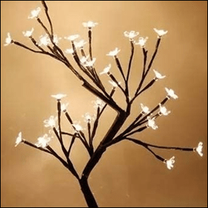 A imagem mostra uma árvore de cerejeira com os galhos pretos e as flores brancas com pequenas luzes de led em seus miolos, que estão acesas. O fundo da imagem é marrom claro.