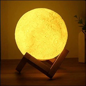 A imagem mostra uma luminária em formato de lua cheia. A lua está acesa com uma luz amarela e está sobre um apoio de madeira. A luminária está em cima de uma mesa de madeira e é possível ver uma parede branca atrás dela.