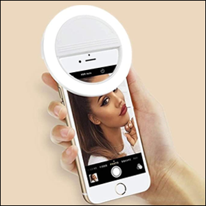A imagem mostra uma mão segurando um celular onde, na parte superior, há um círculo branco com luz led preso ao aparelho. Na tela do celular é possível ver a face de uma mulher. O fundo da imagem é rosa claro.