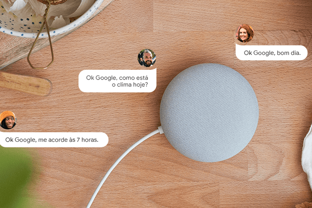 A imagem mostra uma mesa de madeira com um Nest Mini, alto falante inteligente, no formato redondo e na cor cinza, no centro da mesa. Ao redor do aparelho, há alguns balões de falas com frases como: "Ok Google, como está o clima hoje?".