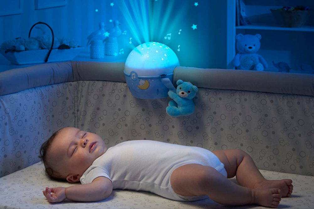 A imagem mostra um bebê dormindo no berço, com um body branco. É possível ver, preso ao berço, o projetor Next to Stars, com uma iluminação azul que ilumina todo o quarto.