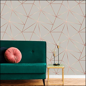 A imagem mostra uma parede com papel de parede com fundo cinza e linhas formando um desenho geométrico em uma cor que imita o efeito de dourado. É possível ver o chão de madeira com um rodapé branco e um sofá de veludo verde escuro, com uma almofada redonda rosa ao lado de uma mesinha quadrada.