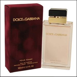 A imagem mostra a caixa do perfume Dolce&Gabbana Pour Femme, aveludada e na cor vinho, com o nome da marca na parte superior em dourado. Ao lado da caixa há o frasco do perfume, de formato retangular, de vidro com um tom rosado. A tampa é prta com a parte inferior em dourado. O fundo da imagem é branco.