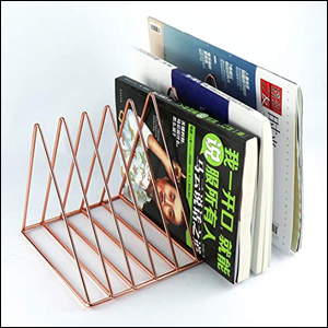 A imagem mostra um porta-arquivos aramado de aço, na cor rose gold, de formato triangular. Entre os triângulos, nos espaços destinados, há algumas revistas e livros. O fundo da imagem é branco.