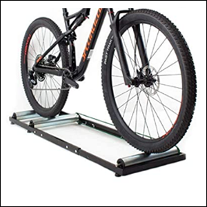 Rolo-Treino-Bike-Triplo-Equilibrio-RT A imagem mostra uma bicicleta em cima do rolo de treino, composto por uma estrutura de metal preta, no meio da qual há 3 rolos de alumínio. O fundo da imagem é branco.