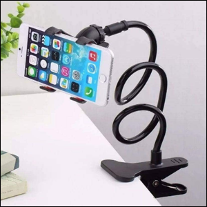 A imagem mostra um suporte para celular preto, preso a uma mesa branca, com seu cabo em zigue-zague e segurando um celular branco, que mostra a tela inicial do iPhone com um fundo azul.