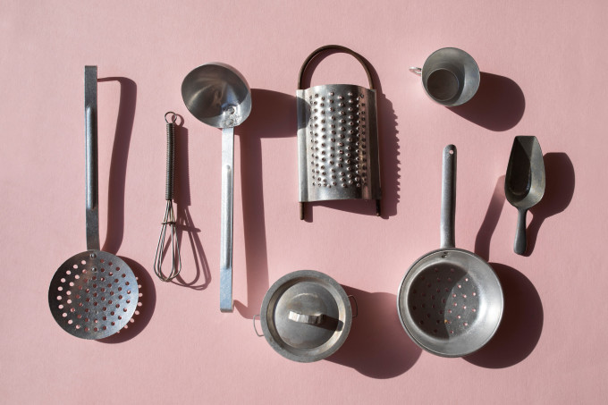 A imagem mostra alguns utensílios de cozinha de aço, como escumadeira, concha, ralador, panela, copo e fouet num fundo rosa.