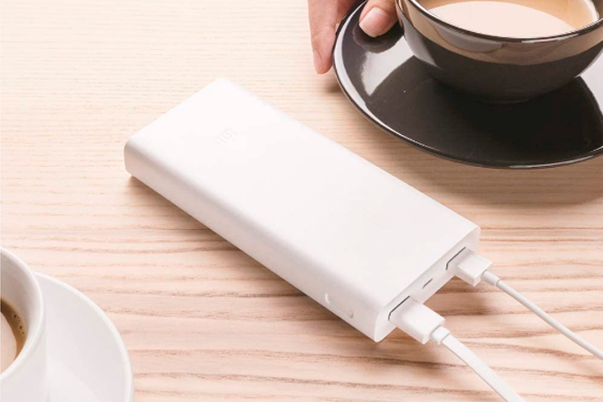A imagem mostra o power bank da Xiaomi, com formato retangular, na cor branco, com dois cabos USB conectados a cada uma de suas saídas. O power bank está sobre uma mesa de madeira e é possível ver o pedaço de duas xícaras de café, uma no canto inferior esquerdo, na cor branca, e outra, de cor marrom, no canto superior direito.