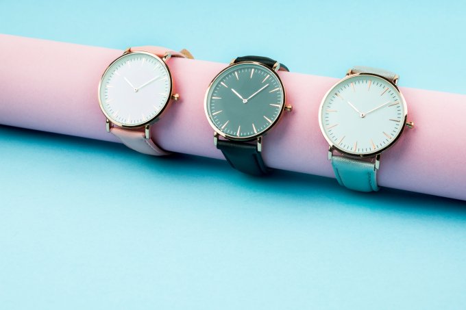 A imagem mostra três relógios em um mostrador da cor rosa, apoiado num fundo azul. O primeiro relógio tem pulseira rosa e visor branco, o segundo tem pulseira preta e visor preto e o último, pulseira azul e visor branco.