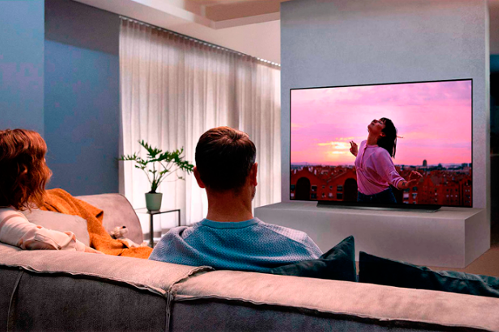 A imagem mostra um casal, sentado de costas para a câmera, num sofá marrom. É possível ver a sala de estar, com paredes azuis, uma janela com cortina brancas do lado esquerdo e um vaso de plantas. Na frente do sofá, há uma tv que mostra uma menina numa paisagem com fundo roxo, por conta do pôr-do-sol.