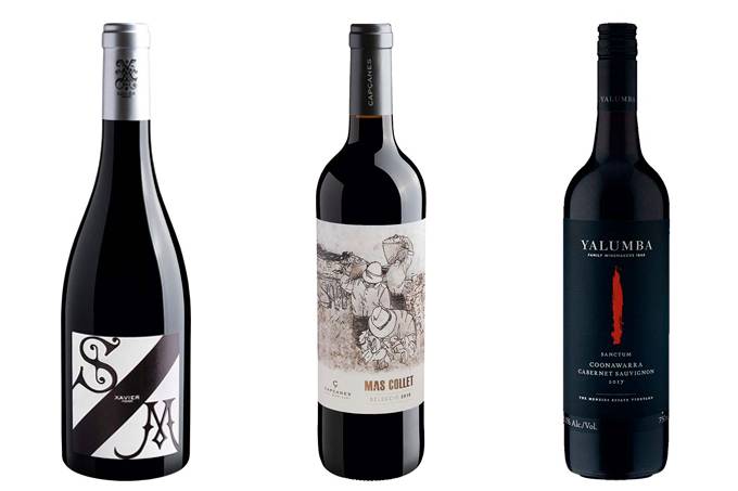 A imagem mostra três garrafas de vinhos diferentes, todas pretas. A primeiro garrafa apresenta um rótulo branco, cortado na diagonal por uma faixa preta, com as letra S e M em preto, em cada lado da faixa. A segunda garrafa possui um rótulo branco com o desenho de alguns homens na colheita, enquanto a última garrafa possui um rótulo preto com um detalhe em vermelho no meio.