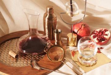 Imagem de objetos de bar: decanter, copo de Whisky, castiçal, coador de drinks, coqueteleira e uma fruta romã de modo decorativo.