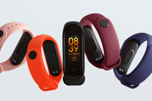 A imagem mostra 5 smartbands, uma de cada cor: rosa, laranja, preta, roxa e azul marinho. Apenas a smartband preta, que está no centro de todas, está virada para frente, dando para ver sua tela, também preta e digital, que mostra o horário em amarelo. O fundo da imagem é cinza.