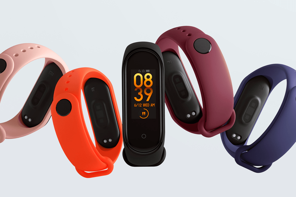 A imagem mostra 5 smartbands, uma de cada cor: rosa, laranja, preta, roxa e azul marinho. Apenas a smartband preta, que está no centro de todas, está virada para frente, dando para ver sua tela, também preta e digital, que mostra o horário em amarelo. O fundo da imagem é cinza.