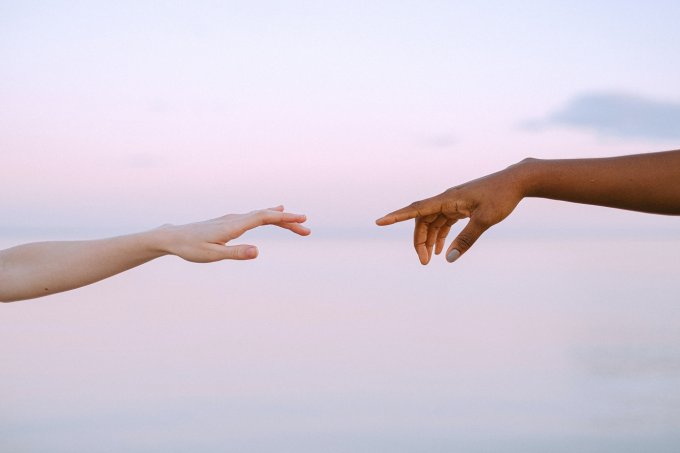 A imagem mostra, do lado esquerdo um braço de uma pessoa branca, com as mãos quase que tocando as mãos de uma pessoa negra, cujo braço está esticado vindo do lado direito da imagem. O fundo da imagem, é um céu azul claro com toques de lilás, como que entardecendo.
