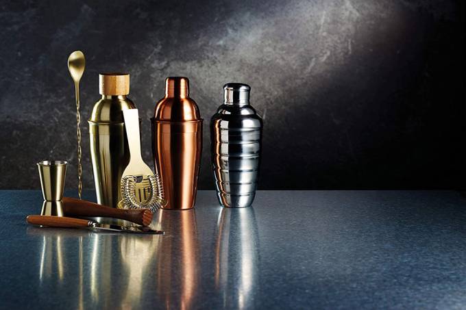 A imagem mostra alguns itens de coquetelaria, nas cores prata, dourado e bronze com alguns detalhes em madeira, num fundo preto e em cima de uma mesa acinzentada.