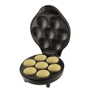 A imagem mostra uma máquina de cupcake no formato redondo, aberta, mostrando seu interior na cor preta com alguns círculos vazados no formato de cupcakes. É possível ver alguns capcakes prontos dentro da máquina. O fundo da imagem é branco.
