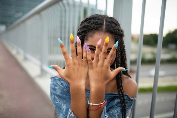 A imagem mostra uma mulher negra, com os cabelos trançados, usando uma blusa azul escura, mostrando suas unhas, pintadas com esmaltes coloridos, para a câmera, em frente ao seu resto.