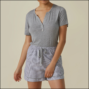 Pijama-Curto-Botões-Amaro A imagem mostra uma mulher negra, do pescoço para baixo, usando uma camiseta cinza com botões na gola e um shorts listrado cinza e branco com amarração na cintura. O fundo da imagem é bege.