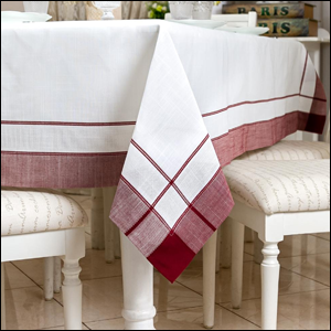 A imagem mostra uma toalha de mesa branca, com as bordas na cor vermelho bordô, sobre uma mesa com as pernas brancas e duas cadeiras ao lado, também brancas com o estofado branco.