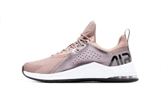 A imagem mostra um pé do tênis da Nike, no modelo Air Max Bella TR 3 feminino, visto de lado, na cor rosa, com a parte da sola branca e alguns detalhes em prata. O fundo da imagem é branco.