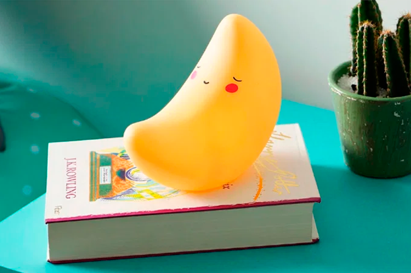 A imagem mostra uma lumin&aacute;ria em formato de lua minguante amarela, com uma cara desenhada, sobre um livro que est&aacute; em cima de uma mesa de cabeceira azul. &Eacute; poss&iacute;vel ver tamb&eacute;m um pequeno vaso com um cacto.