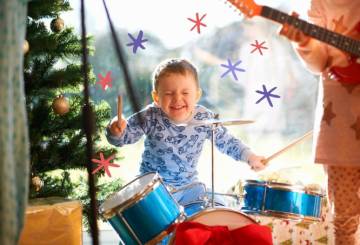 A imagem mostra um menino, de pele branca, tocando um bateria na cor azul, com um pijama na cor azul claro. Ele está cercado por pequenas estrelas desenhadas nas cores roxo e rosa. É possível ver parte de uma árvore da Natal ao seu lado esquerdo e uma parte de outra criança, de pijama rosa, tocando guitarra, ao seu lado direito.