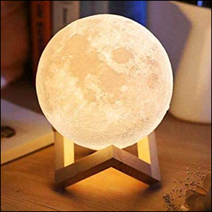 A imagem mostra uma luminária em formato de lua, iluminada, sobre um suporte de madeira, sobre uma mesa de madeira.