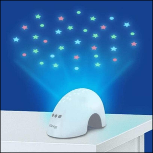 A imagem mostra uma luminária em formato de semi-círculo, na cor branca, apoiado em uma mesa branca e projetando luzes em formato de estrelas e bolinhas numa parede azul escura.