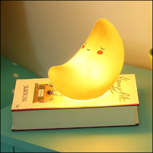 A imagem mostra uma luminária em formato de lua minguante, amarela, com uma carinha desenhada, acesa sobre um livro que está sobre uma mesa de cabeceira da cor azul.