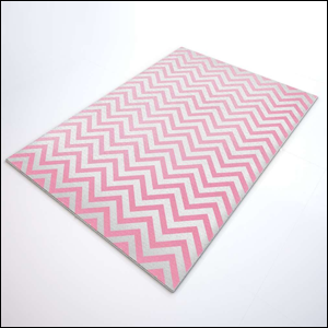 A imagem mostra um tapete retangular, em estampa de chevron, zigue e zague, nas cores branco e rosa. O fundo da imagem é branco.