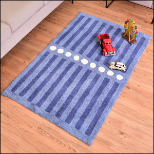 A imagem mostra um tapete retangular de algodão, na cor azul com listras azul escuro, separadas no meio por uma fila de círculos brancos alinhados. Ele está sobre um piso de madeira e, sobre ele, há um carrinho de brinquedo vermelho e uma roda gigante de madeira de brinquedo.