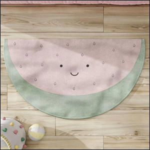 A imagem mostra um tapete em formato de melancia, nas cores verde claro e rosa claro. A melancia possui pequenos pontinhos pretos e uma carinha desenhada, e está sobre um chão de madeira clara.