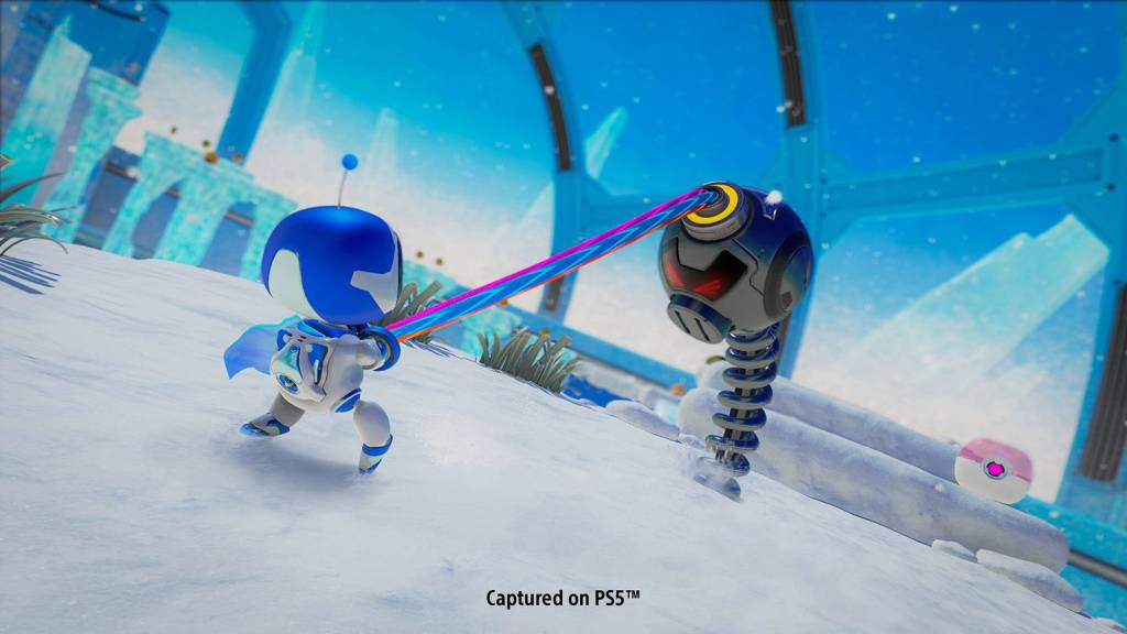 astro-playroom A imagem mostra uma cena do jogo Astro's Playroom, na qual o personagem, um robô nas cores branco e azul, está batalhando com um robô maior, da cor cinza e com o corpo em formato de mola. O fundo do jogo é azul e o chão parece estar coberto de neve.
