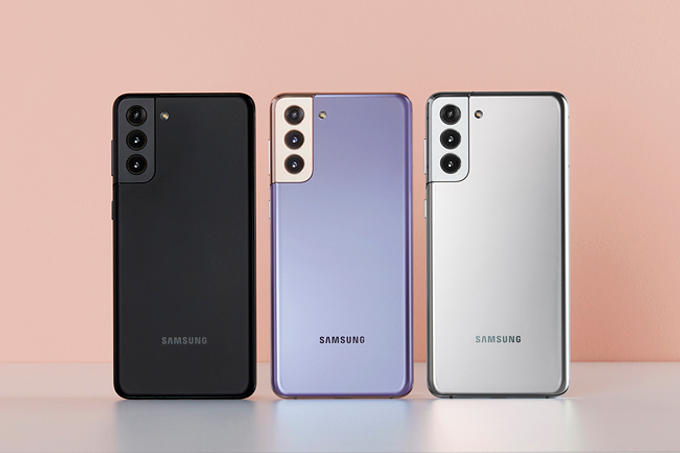 A imagem mostra três celulares Galaxy S21 com a parte traseira para frente, cada um de uma cor, um preto, outro lilás e outro prata, em pé sobre uma mesa branca e com o fundo na cor rosa claro.
