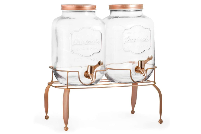 A imagem mostra uma suqueira, com duas jarras de vidro com tampas em rose gold e torneiras da mesma cor, sobre um suporte de metal também rose gold.