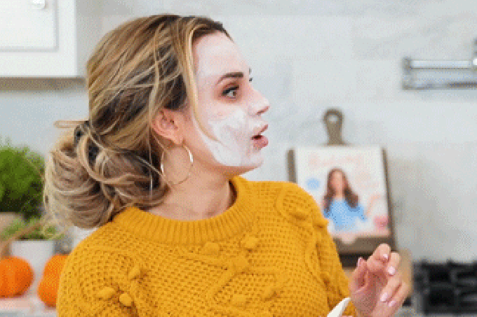 A imagem é um gif animado de uma mulher branca, com os cabelos loiros presos num coque. Ela está usando um suéter da cor amarelo mostarda. Ela está com o rosto coberto por um creme branco, enquanto segura uma embalagem na mão, enquanto fala "Well...", como pode ser visto na legenda da imagem, em letras brancas.