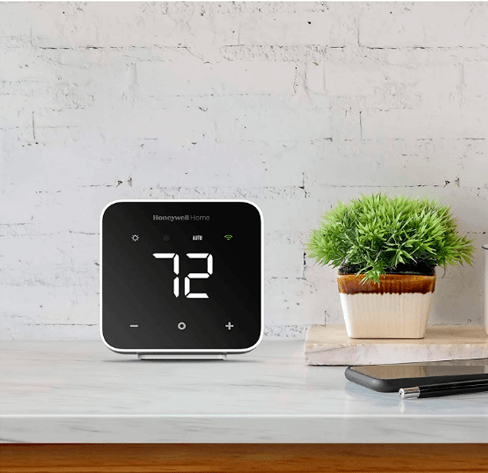 A imagem mostra um balcão branco com um termostato quadrado preto em sua superfície, ao lado de um celular e um pequeno vaso de planta.