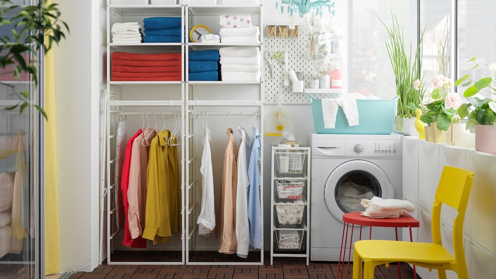 A imagem mostra uma lavanderia, com máquina de lavar roupas, uma estante com cabides e roupas penduradas e uma cadeira amarela.