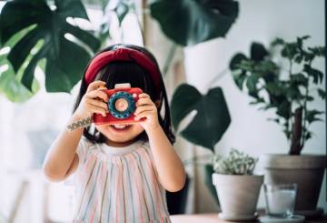 A imagem mostra uma criança branca, com os cabelos escuros e uma franja reta, com uma faixa vermelha no cabelo, segurando uma câmera fotográfica de brinquedo em frente ao rosto. Ela está com um vestido listrado e, atrás dela, é possível ver algumas plantas.