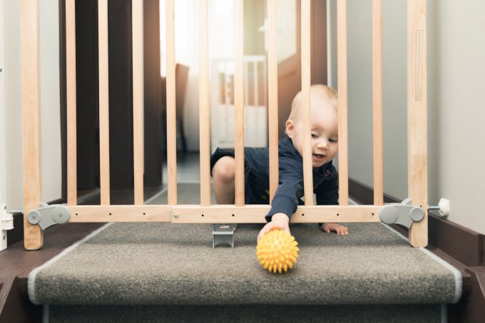 A imagem mostra um bebê atrás de uma cerca de proteção de madeira tentando pegar uma bola amarela do outro lado da cerca.