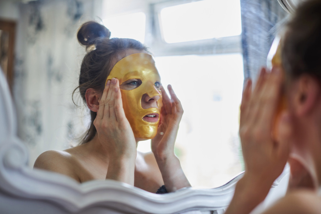 GettyImages-928890656 Mulher com máscara facial dourada se olhando no espelho.