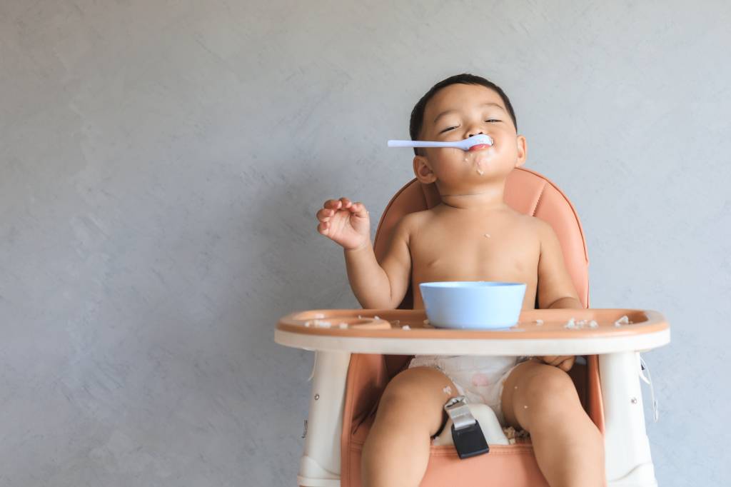 A imagem mostra um bebê asiático sentado em uma cadeira de alimentação, com uma colher na boca. O fundo da imagem é cinza.
