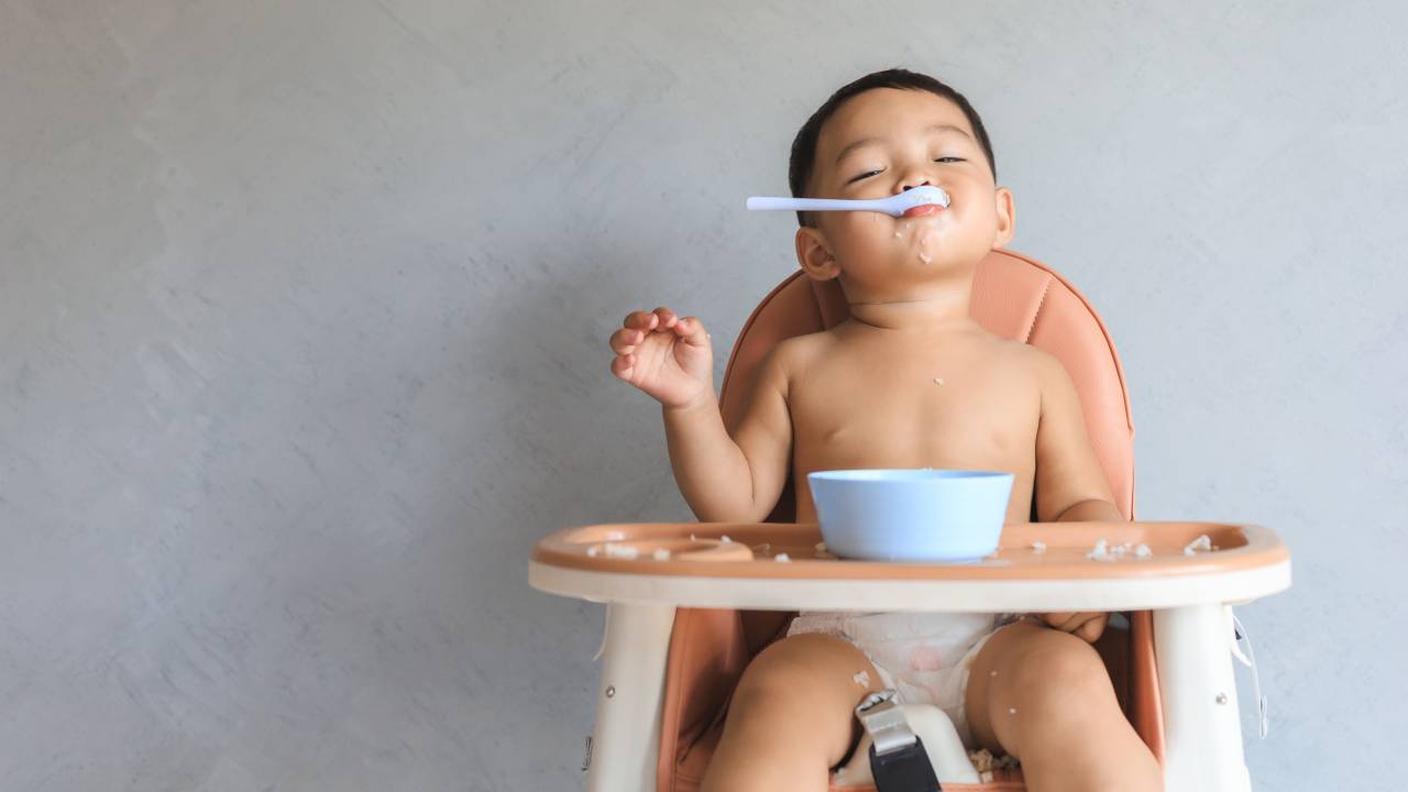 A imagem mostra um bebê asiático sentado em uma cadeira de alimentação, com uma colher na boca. O fundo da imagem é cinza.