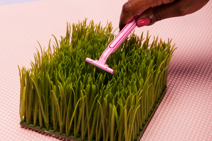 A imagem mostra a mão de uma mulher negra segurando uma lâmina de depilação na cor rosa em cima de um quadrado de grama verde num fundo rosa.