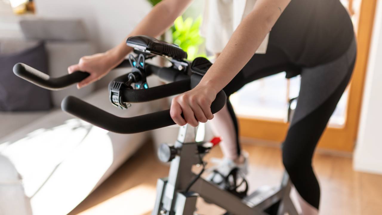A imagem mostra uma pessoa pedalando em uma bicicleta ergométrica na sala de sua casa.